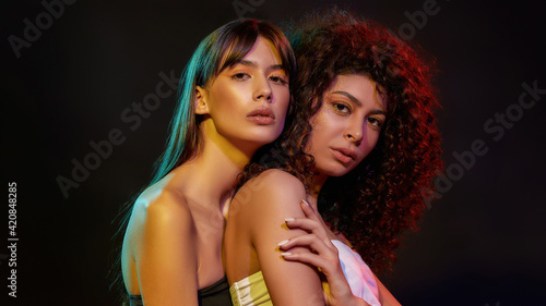 Two gorgeous young brunette women with professional art makeup posing together in neon light isolated over black background