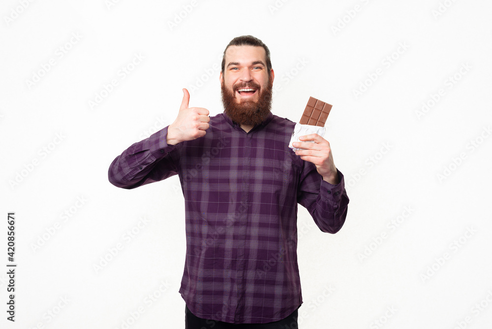 Portrait of young handsome bearded man showing thumb up and holding bar of chocolate Portrait of young handsome bearded man showing thumb up and holding bar of chocolate