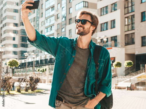 Handsome smiling stylish hipster lambersexual model.Man dressed in green shirt.Male posing in the street at skyscraper background in sunglasses. Outdoors at sunset with backpack. Taking selfie