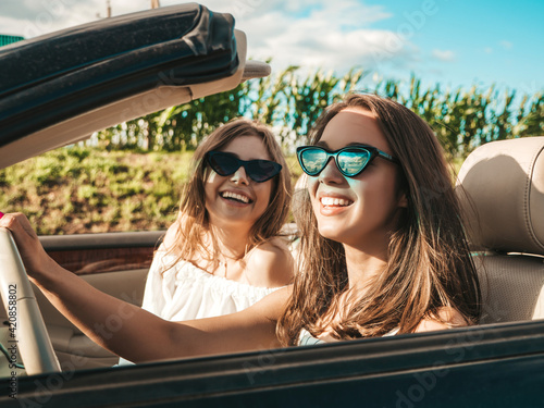 Portrait of two young beautiful and smiling hipster female in convertible car. Sexy carefree women driving cabriolet. Positive models riding and having fun in sunglasses outdoors.Enjoying summer days © halayalex