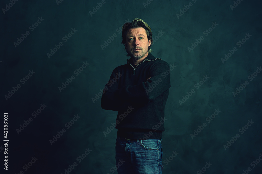 Middle aged man with stubble beard in green woolen sweater and jeans in front of grey wall.