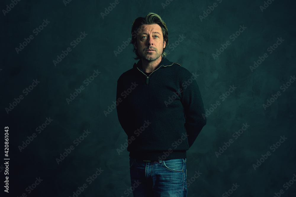 Middle aged man with stubble beard in green woolen sweater and jeans in front of grey wall.