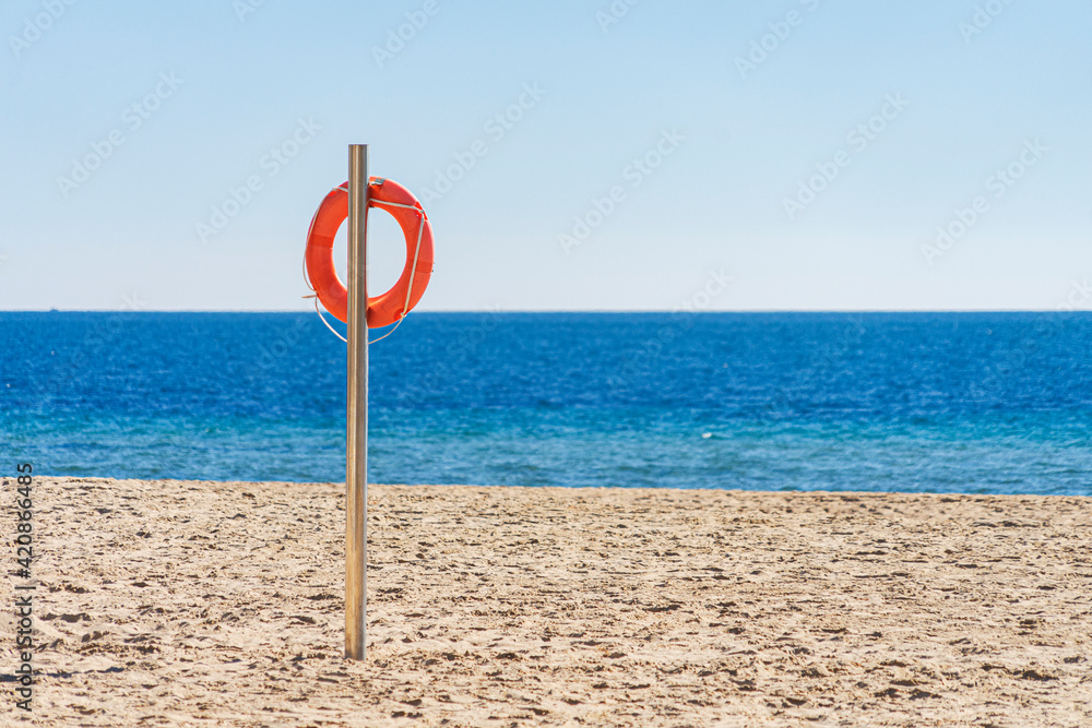 Orange lifebuoy in empty sandy beach in front of Mediterranean sea. Life saving lifeguard buoy, lifebelt on the pole