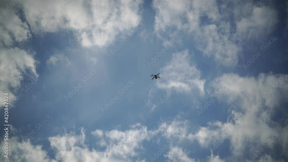 The drone flies against a background of blue sky and clouds, a view from below. The quadcopter with the camera hovers at a low altitude. Fast-rotating drone propellers.