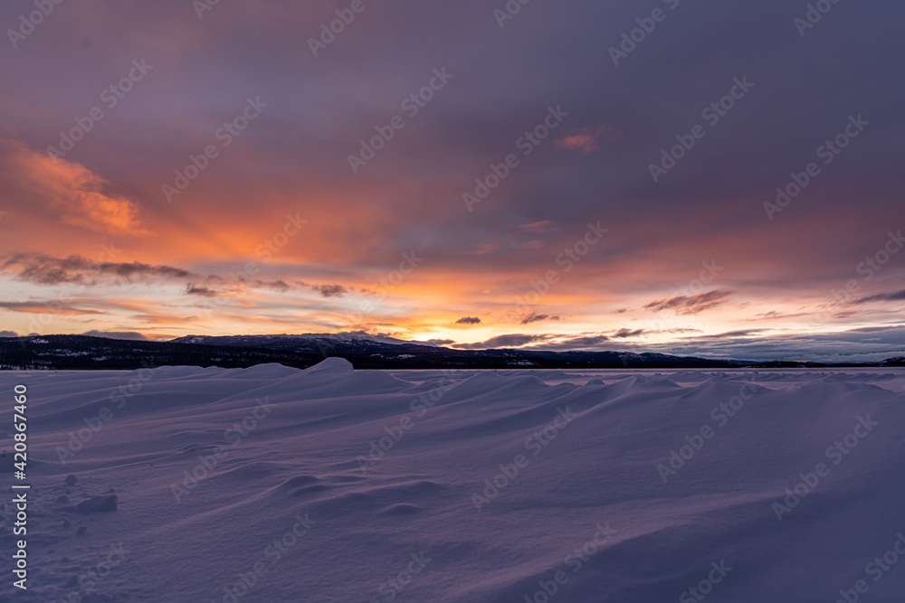 Winter frozen lake scene in northern Canada on a stunning cloudy sunset afternoon in March with white snow, mountains in background and iconic Canadian landscape in the north. 