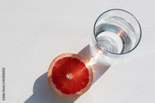 Summer still life with glass of water and grapefruit slice on white background.  Sunny day. Hard light, shadow
