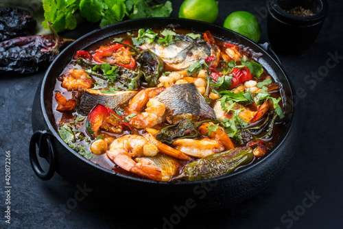 Modern style traditional Mexican seafood pozole soup with fish, king prawns and hominy in a clear sauce served as close-up in design pot