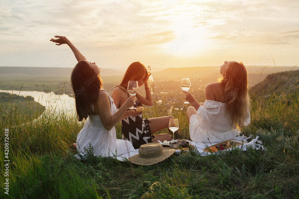 Three young happy girlfriends in elegant dresses is having picnic on the hill on sunset.