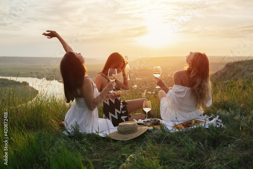 Three young happy girlfriends in elegant dresses is having picnic on the hill on sunset.