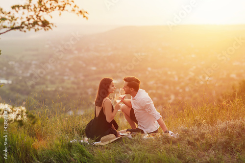 Attractive couple in love enjoying picnic on the hill at sunset. Happy young couple making a toast with white wine. Romance, dating and love concept.