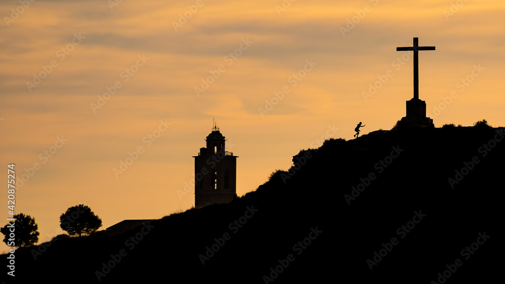 silhouette of a boy jumping and running on a hilltop with a large cross and a church in the background