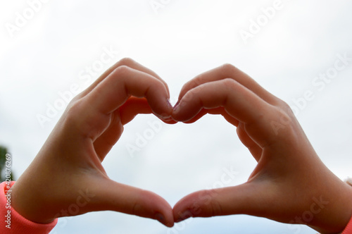 the girl s hands folded the shape of a heart against a gray sky  the heart is a symbol of love  devotion and care