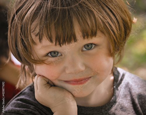 portrait of a little child with blue eyes and bang-cut hair. Grandaughter at her sister's softball game in Windsor in Broome County in Upstate NY.  Wide-eyed and an ornery smile says it all.	