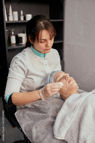 A female cosmetologist cleans the face of a young beautiful female client with cotton pads. Professional beauty salon. Vertical photo.
