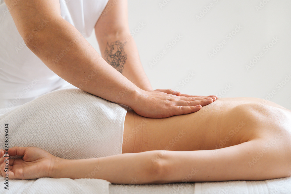 Side view of male masseur massaging small of back of young woman lying on massage table at light spa salon. Experienced chiropractor performs wellness treatments for lady with back pain.