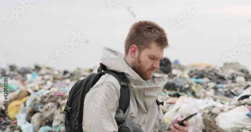 A working man against the backdrop of burning garbage. A lot of plastic bags thrown to the dump. From the plastic debris goes smoke. On the man, the khaki color encephalic suit photo