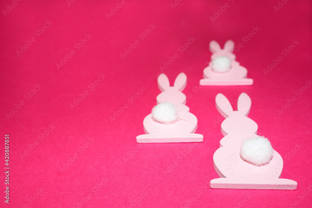 On the right, on a pink background, three pink wooden figures of an Easter bunny with a white tail lie on the side . Easter Symbol On the right, on a pink background, three pink wooden figures of an Easter bunny with a white tail lie on the side . Easter Symbol