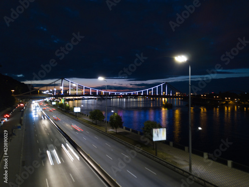 Night filming of the embankment darts by the water, cars go on the road, against the backdrop of a beautiful pedestrian bridge
