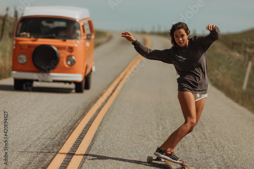 Young female skateboarder swerve skateboarding down rural road, followed by recreational vehicle, Jalama, California, USA photo