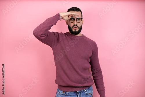 Handsome man wearing glasses and casual clothes over pink background very happy and smiling looking far away with hand over head. searching concept.