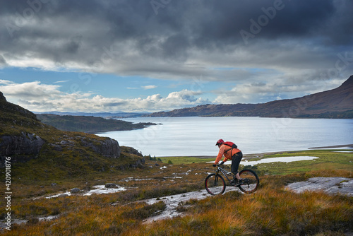 Male mountain biker biking down toward loch in mountain landscape,  Achnasheen, Scottish Highlands, Scotland