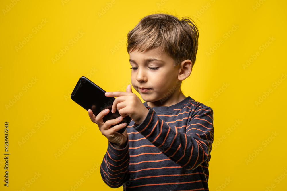 Front view portrait of small caucasian boy holding a smartphone in front of yellow background - little kid using mobile phone to take photos or play games while standing in studio with copy space Front view portrait of small caucasian boy holding a smartphone in front of yellow background - little kid using mobile phone to take photos or play games while standing in studio with copy space