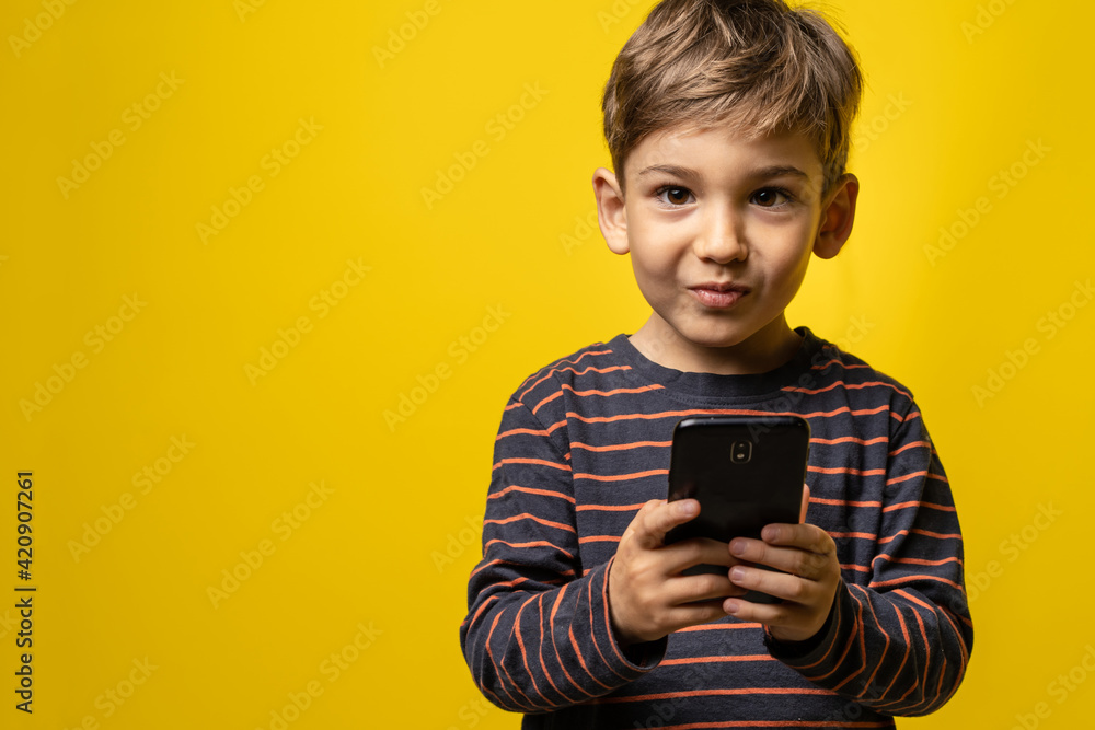 Front view portrait of small caucasian boy holding a smartphone in front of yellow background - little kid using mobile phone to take photos or play games while standing in studio with copy space Front view portrait of small caucasian boy holding a smartphone in front of yellow background - little kid using mobile phone to take photos or play games while standing in studio with copy space