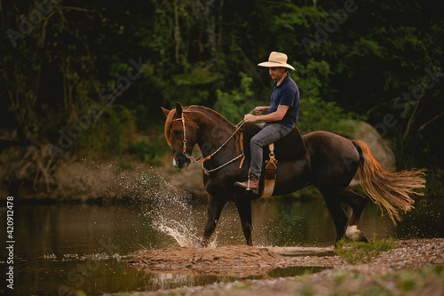 White man riding a horse inside a river, wearing a straw hat, with his horse in a saddle and throwing water into the air.