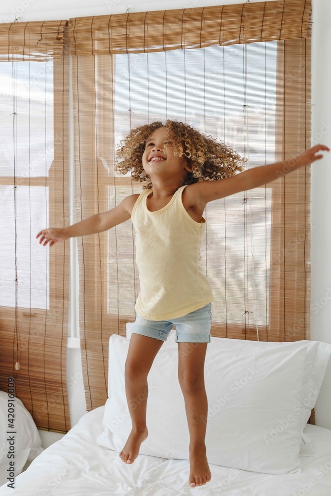 Little girl jumping with arms open on bed in beach house Little girl jumping with arms open on bed in beach house