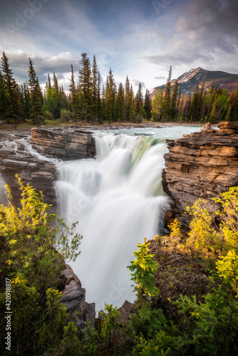 Athabasca Falls in Jasper National Park in the evening as the setting sun shines on the mountains  cliffs  and greens.