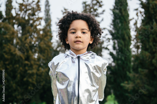 Black boy wearing a silver clothing, looking at the camera. In a park background. Copyspace. Kids, astronauts, extraterrestrials and black people concept