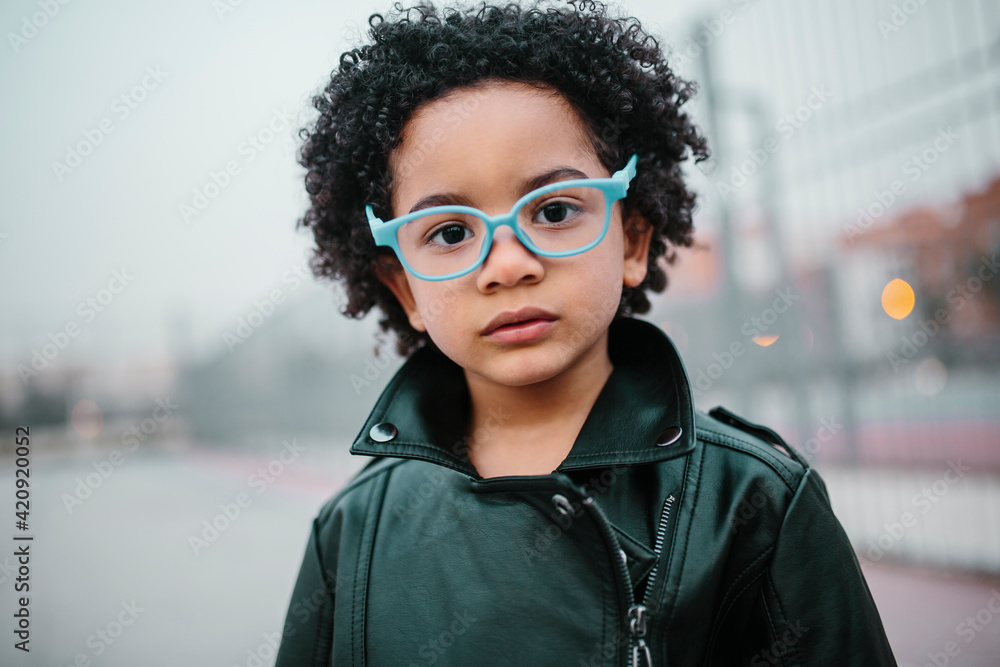 Portrait of a afro child with blue glasses, looking at the camera, with a serious expression. In a courts background. Kids, smartphones and black people concept.