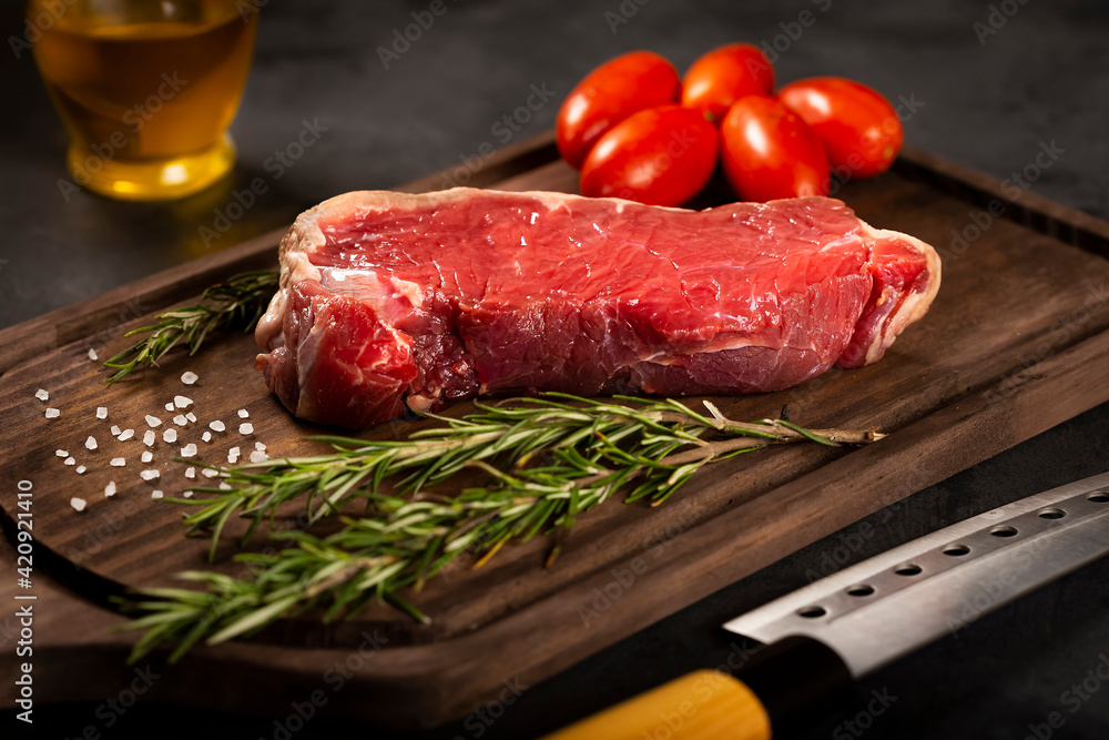 Fresh raw steak on wooden board on a dark background with salt, tomatoes and rosemary.
