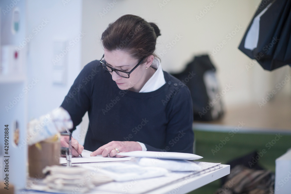 Female tailor wearing glasses, sitting at table, writing. Female tailor wearing glasses, sitting at table, writing.