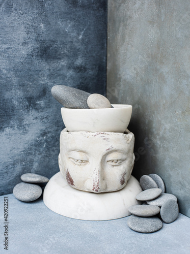 A pestle and mortar standing on a model of a head beside grey pebbles in a grey tiled corner. photo