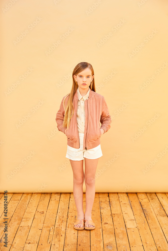 Portrait of girl with long blond hair wearing shorts, shirt and pink jacket, on pale yellow background. Portrait of girl with long blond hair wearing shorts, shirt and pink jacket, on pale yellow background.