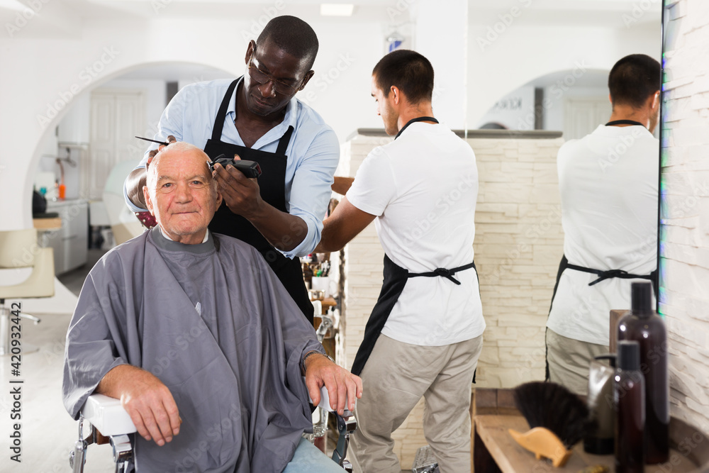 Elderly man getting haircutting with electric clipper from professional African barber in salon