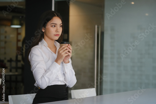 Young woman office worker holding coffee cup or tea and looking away thoughtfully.