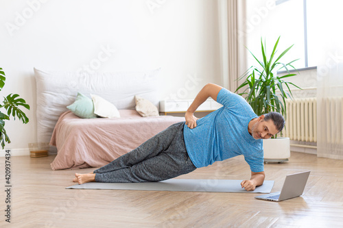 Middle-aged man in front of laptop monitor doing a side plank exercise. He follows the guidance of his online coach.