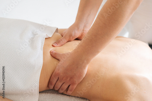 Close-up hands of male masseur massaging small of back of young woman lying on massage table at light spa salon. Experienced chiropractor performs wellness treatments for lady with back pain.