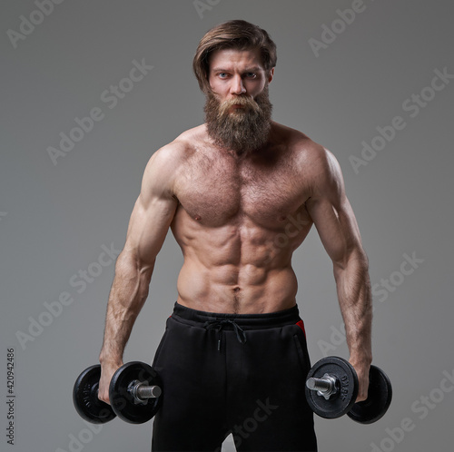 Brutal guy with muscular build posing in gray background with dumbells
