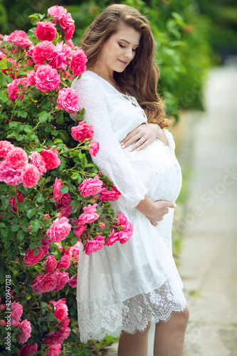 Smiling pregnant woman resting in near roses. Posing outdoors. Motherhood. High quality photo.