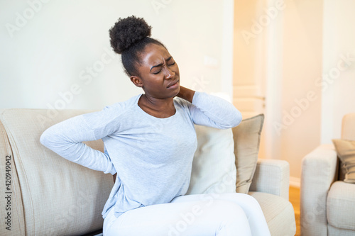 Hurt african American young woman sitting on sofa touching back suffering from painful backache, unwell black millennial girl awake from sleep having strong spinal muscular spasm strain