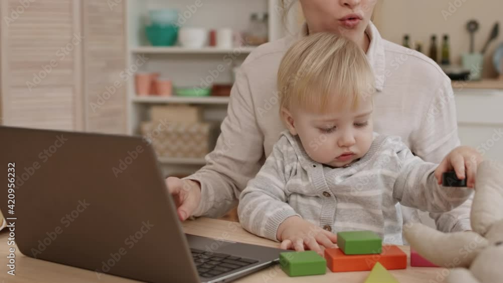 Blond-haired blue-eyed Caucasian toddler playing with colorful toy, sitting in arms of busy cropped parent, who trying working on laptop computer at home Blond-haired blue-eyed Caucasian toddler playing with colorful toy, sitting in arms of busy cropped parent, who trying working on laptop computer at home