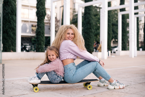 Mother and daughter sit on skateboard. Woman with unusual, lush and long hair. Small girl and woman smiles and happy. Same colour of clothes, get fun outdoor in weekend. Small kid study skating. 