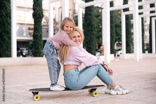 Mother and daughter sit on skateboard. Woman with unusual, lush and long hair. Small girl and woman smiles and happy. same colour of clothes, get fun outdoor in weekend. Small kid study skating. 