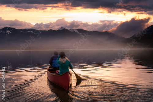 Couple friends canoeing on a wooden canoe during a colorful sunny sunset. Cloudy Sky Artistic Render. Taken in Harrison River, East of Vancouver, British Columbia, Canada. © edb3_16