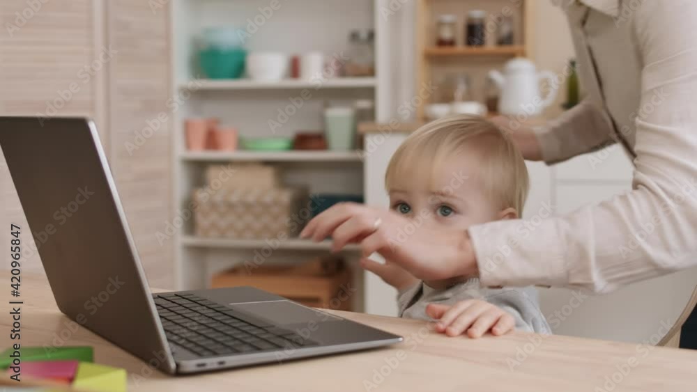 Chest-up of cute blond-haired Caucasian toddler sitting by desk, pressing on keys of laptop, unrecognizable parent checking on child, boy resuming playing with laptop, having fun, smiling Chest-up of cute blond-haired Caucasian toddler sitting by desk, pressing on keys of laptop, unrecognizable parent checking on child, boy resuming playing with laptop, having fun, smiling