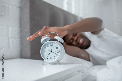A young man in white tshirt lying in bed and feeling annoyed by the sound of alarm-clock