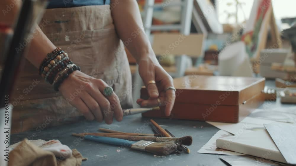 Close up shot of hands of unrecognizable female artist choosing paintbrush on desk while preparing for drawing in studio Close up shot of hands of unrecognizable female artist choosing paintbrush on desk while preparing for drawing in studio